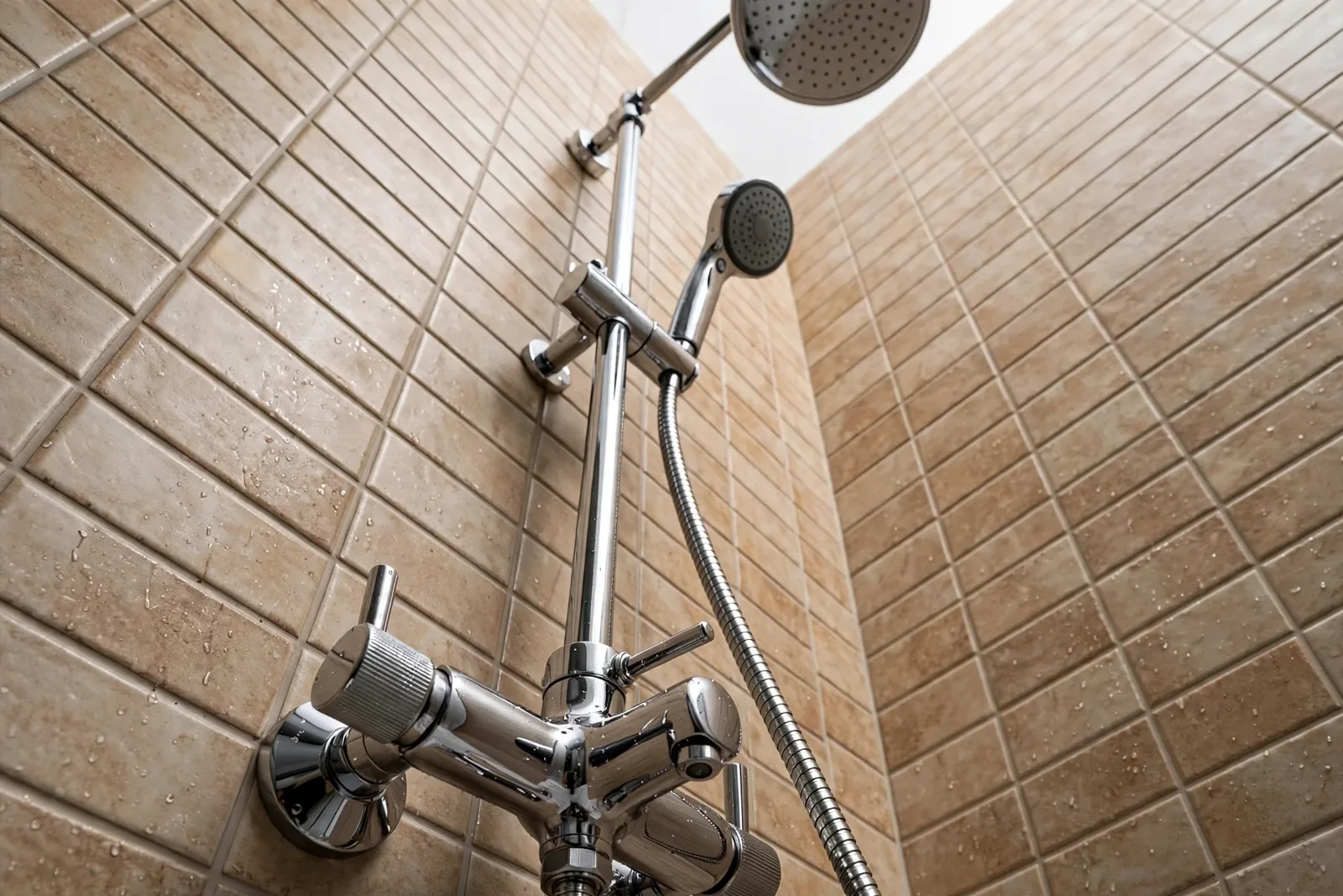 Chromowany natynkowy zestaw prysznicowy z baterią termostatyczną, wylewką, wężem, słuchawką i deszczownicą na tle beżowych płytek.
